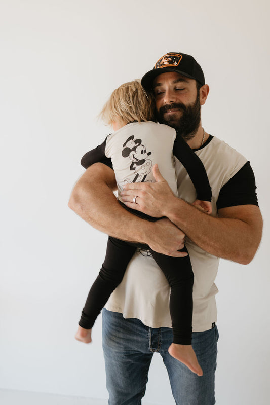 A bearded man wearing a baseball cap lovingly holds a young child, who is dressed in "Steamboat Willie" Bamboo Two Piece Pajamas by forever french baby. They are casually dressed in hypo-allergenic clothing, set against a minimalistic backdrop with a plain, light-colored background.