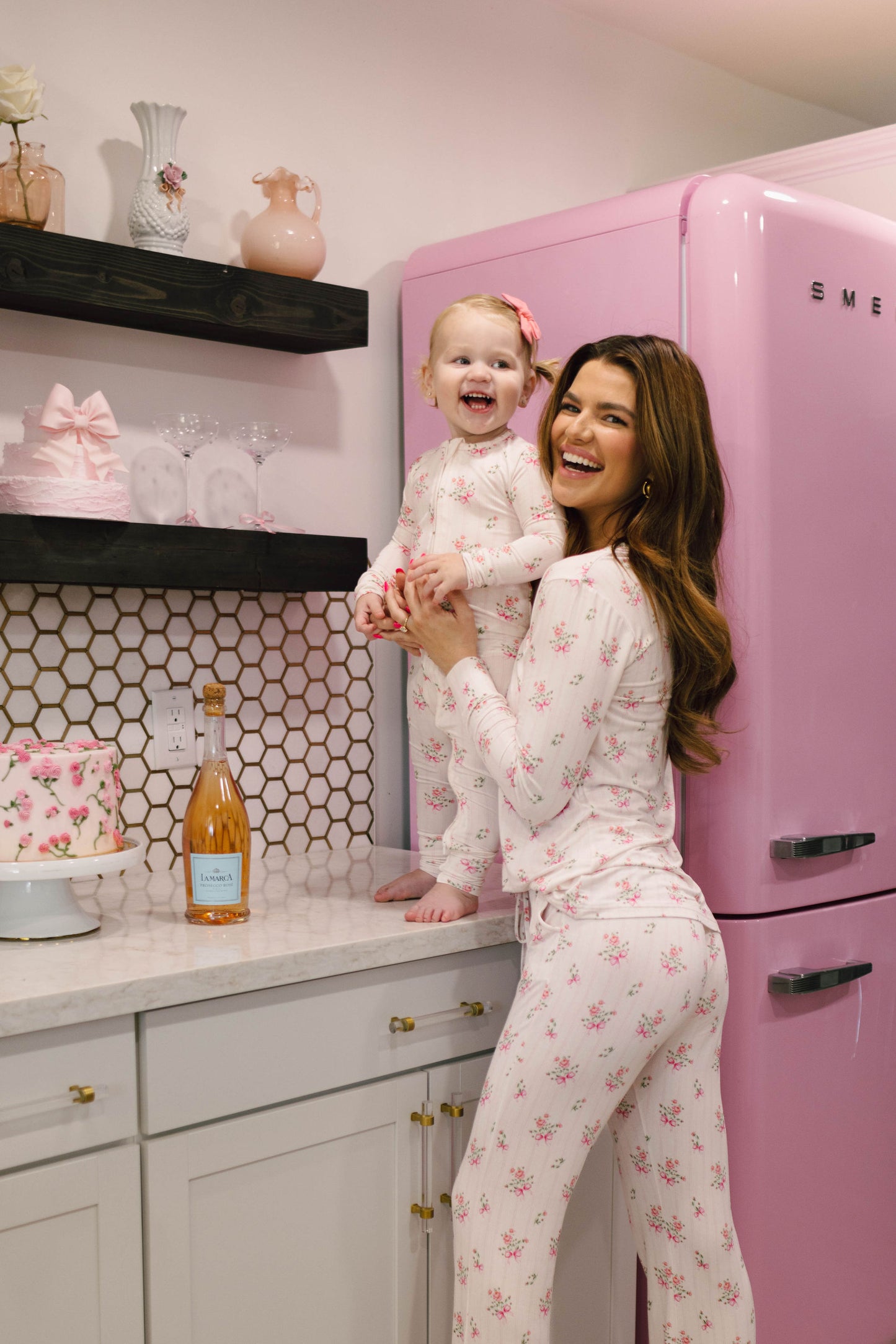 A woman and a child, both dressed in matching Kendy 2 Women's Bamboo Pajamas from the Kendy Floral collection, stand in a light-colored kitchen with a pink fridge. The woman holds the smiling child who is standing on the counter. Nearby, there's a pink cake and a bottle of rosé wine.