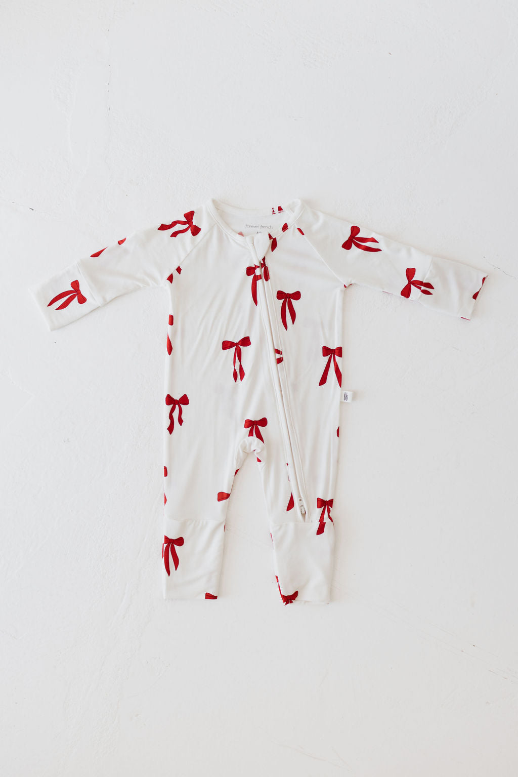 The Bamboo Zip Pajamas | Ribbons & Bows by forever french baby is a white onesie designed for babies, featuring long sleeves and a charming pattern of red bows. Made from hypo-allergenic material to protect delicate skin, the onesie displays its front and convenient zip closure when laid flat on a light surface.