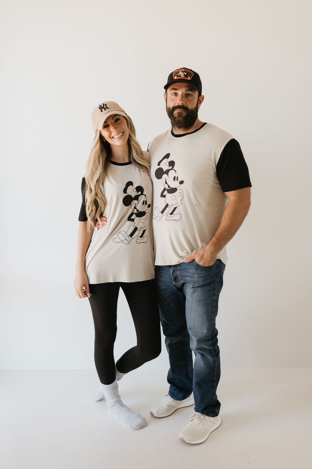 A woman and a man stand in casual attire against a plain background, wearing matching Adult Bamboo Tees with Steamboat Willie designs from forever french baby. The woman pairs hers with leggings and a cap, while the man chooses jeans and a cap. They smile warmly as he wraps his arm around her.