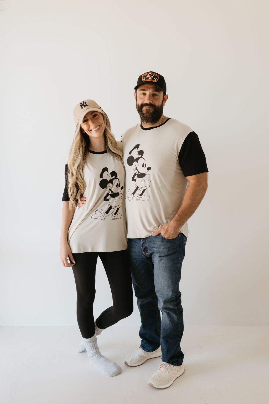 A smiling couple stands together in matching casual outfits from forever french baby, each sporting the Adult Bamboo Tee with a delightful Steamboat Willie motif. The woman complements her look with a light cap and black leggings, while the man pairs his tee with jeans and a dark cap, as they pose against a plain white background.