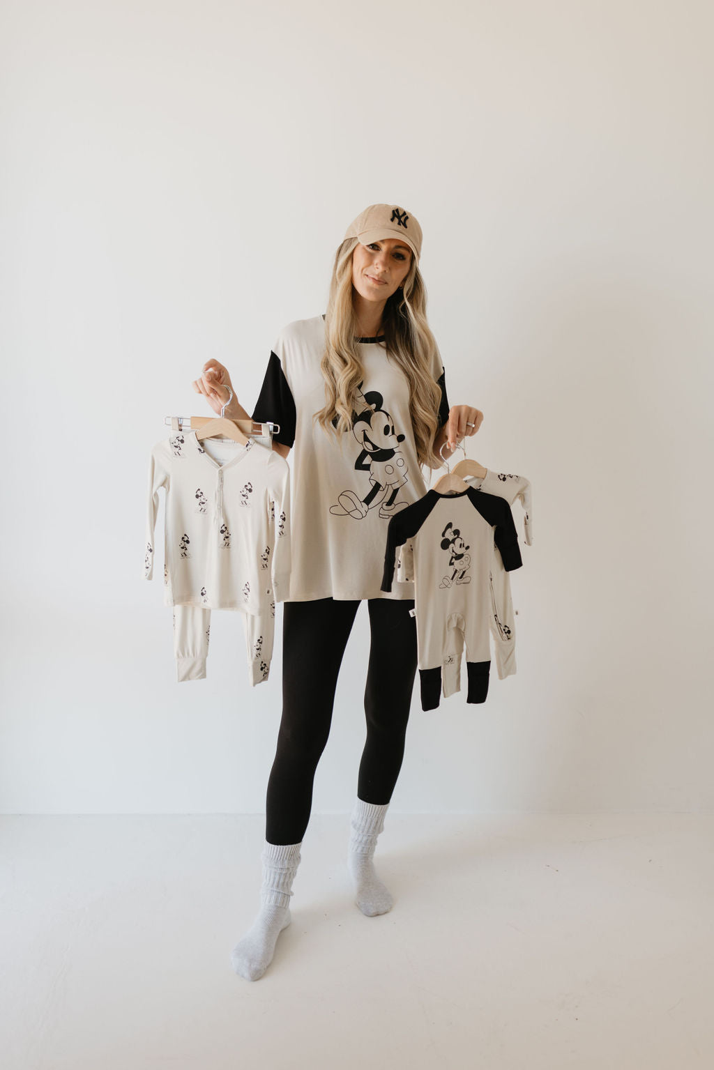 A person with long hair, wearing a baseball cap, an oversized Mickey Mouse shirt, black leggings, and socks, holds up two matching children's Bamboo Zip Pajamas from forever french baby featuring hypo-allergenic fabric against a plain background.