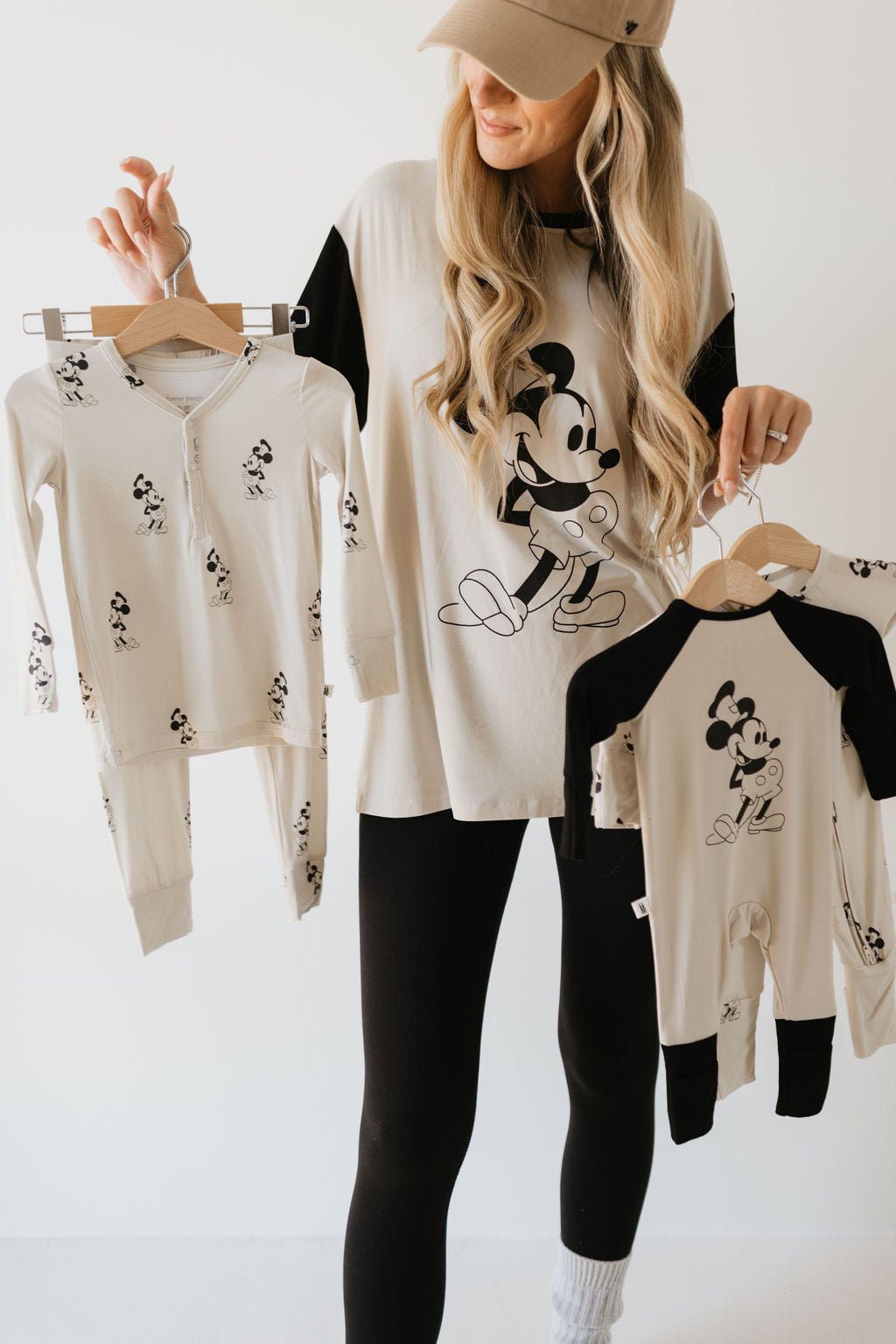 A woman showcases matching Disney-themed pajamas in neutral tones featuring a classic Steamboat Willie design. She wears a comfy Adult Bamboo Tee from forever french baby, paired with black leggings and a beige cap over her long blonde hair. Her infectious smile lights up against the white background.