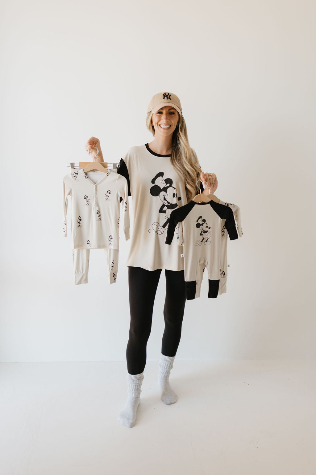 A person in a beige cap and black leggings is smiling while holding up matching Mickey Mouse pajamas. They are wearing the "Adult Bamboo Tee | Steamboat Willie" by forever french baby, standing on a white background.