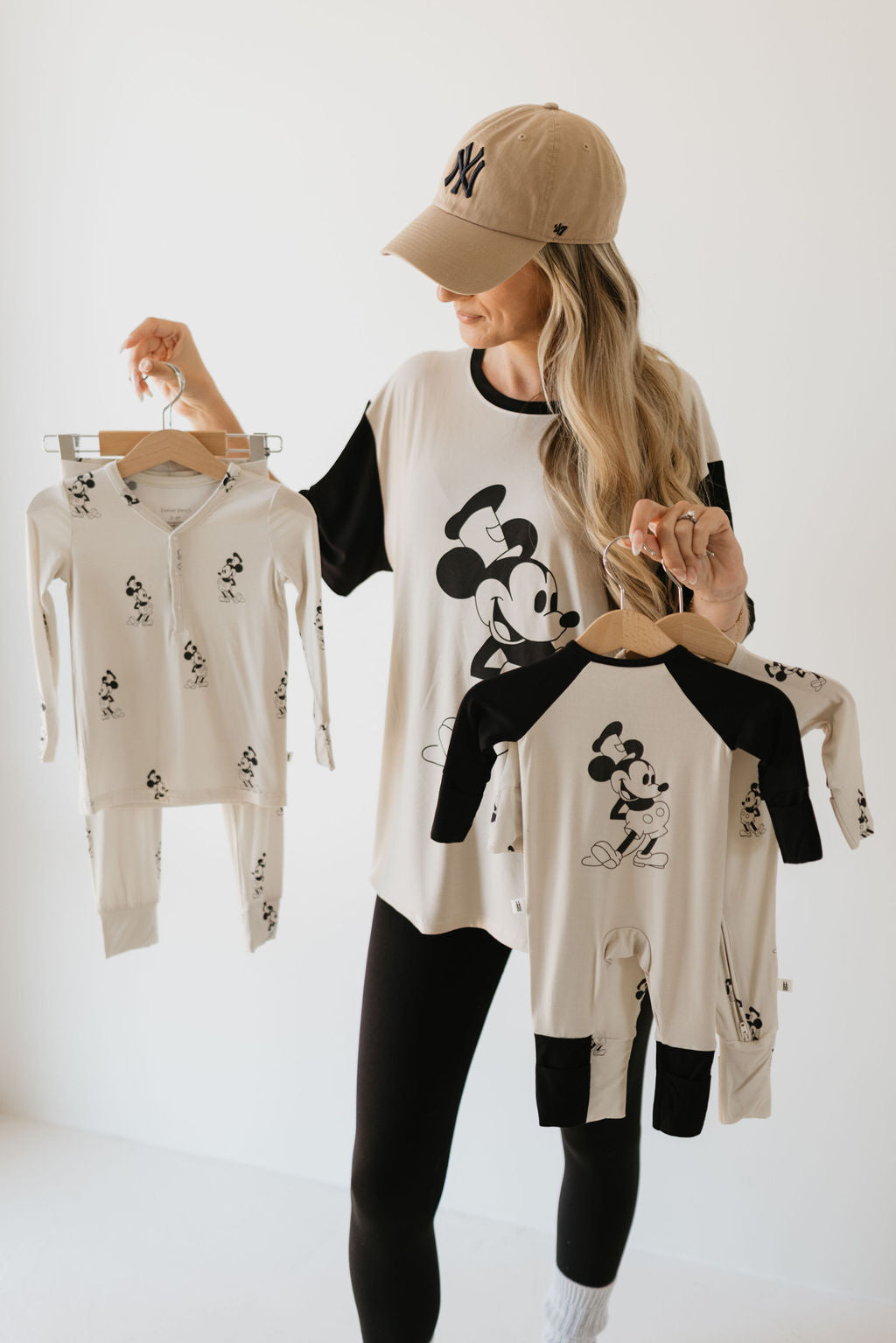 A person in an Adult Bamboo Tee featuring Steamboat Willie from forever french baby holds two Steamboat Willie-themed baby outfits on hangers. They are wearing a beige baseball cap and black leggings, standing against a plain white background.