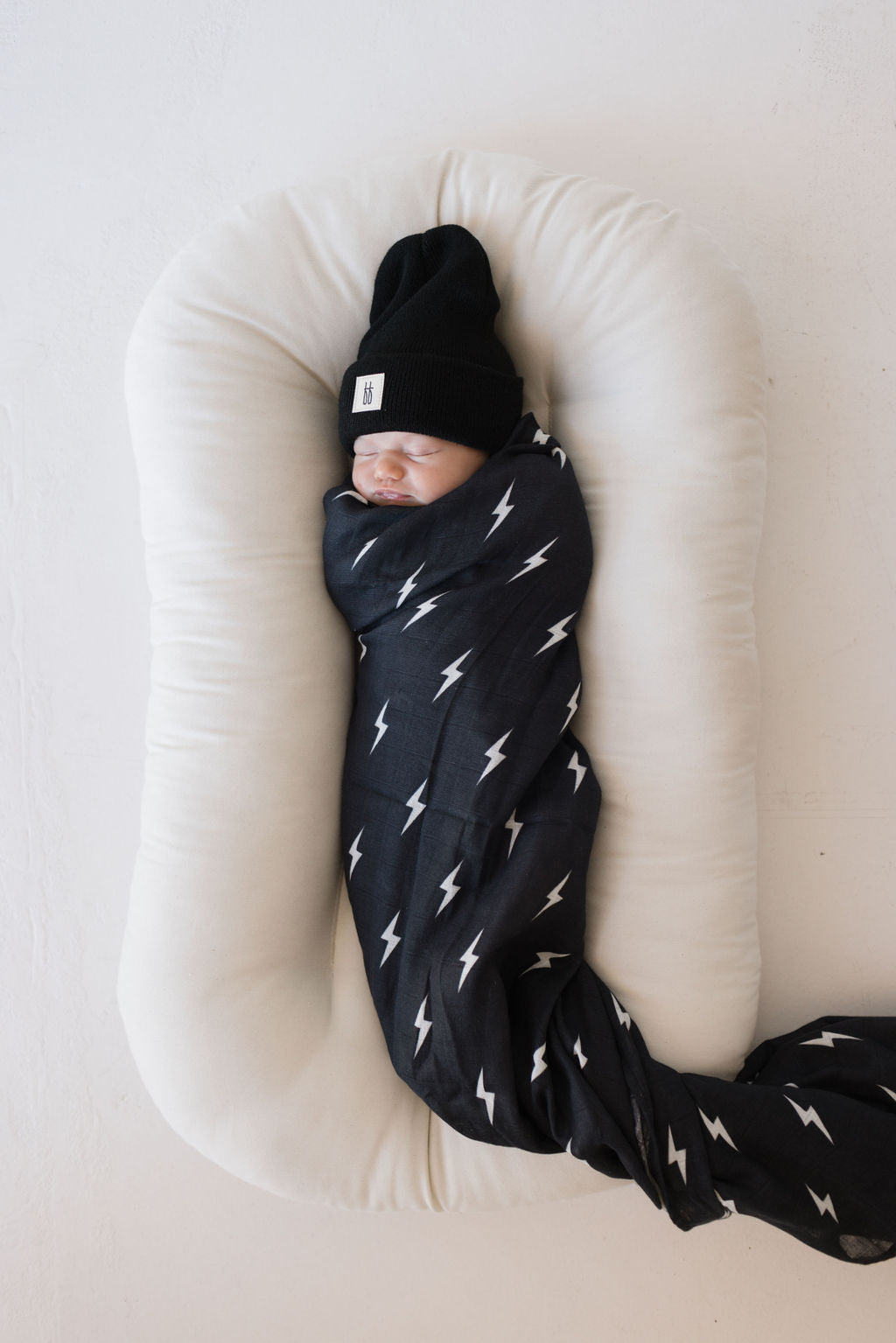 A baby is swaddled in the "Muslin Swaddle | Midnight Lightning Bolt" by forever french baby, lying on a plush white cushion. The baby is wearing a black beanie with a small tag on it, surrounded by a light, neutral background.