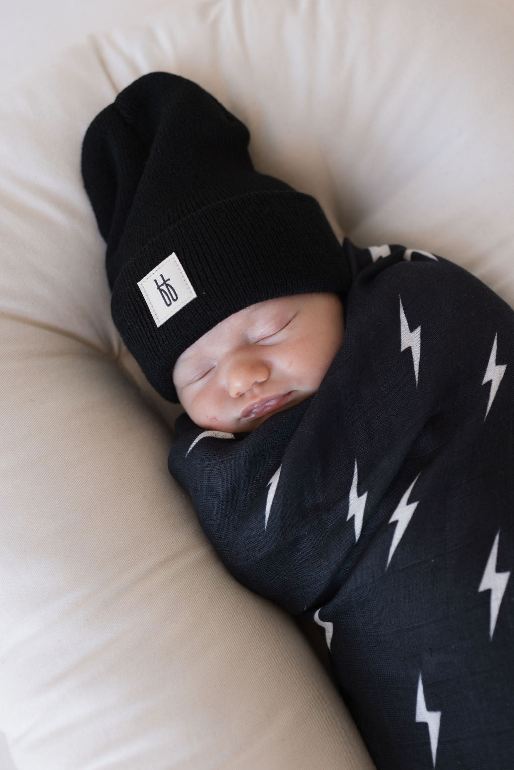 A sleeping baby is swaddled in the Muslin Swaddle | Midnight Lightning Bolt by forever french baby, featuring a black design adorned with white lightning bolt patterns. The baby is also wearing a black knit hat with a small tag and lies peacefully on a cushioned surface.