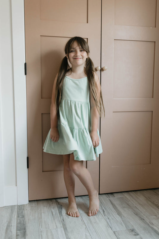 A young girl with long hair stands barefoot in front of a light brown door, smiling and wearing the Children's Dress | Vintage Washed Spearmint by forever french baby. She has her arms relaxed at her sides and one knee slightly bent, with a light wood-colored floor beneath her.