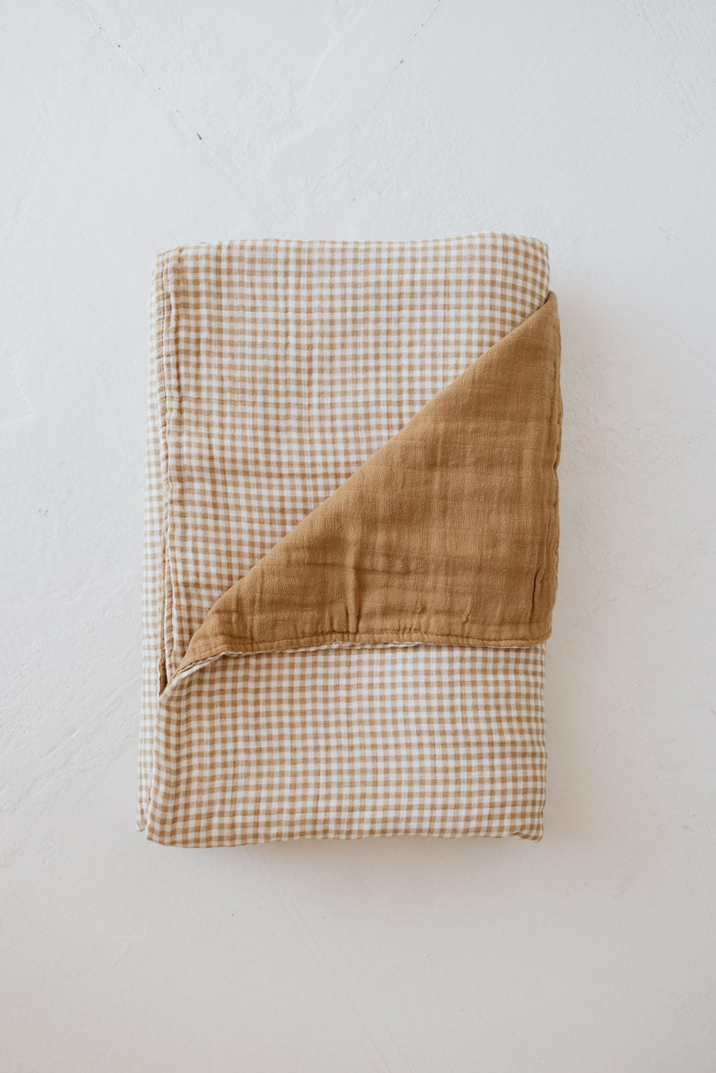 The Muslin Quilt | Harvest Days by forever french baby features a folded checkered blanket with one corner flipped, revealing a solid brown underside. It's crafted from soft muslin cotton and showcases a gingham quilt pattern in beige and white against a light background, making it an elegant addition that brings timeless charm to any space.