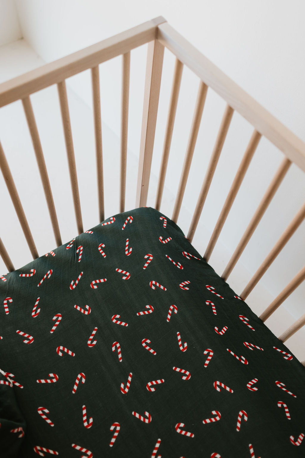 A wooden crib with a Muslin Crib Sheet in the Candy Cane Lane pattern from forever french baby is set against a white wall in a minimalist room.