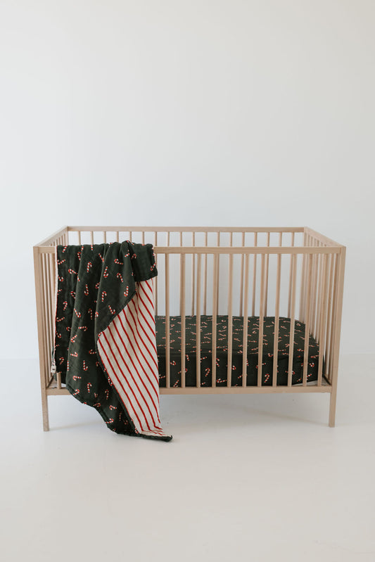 A minimalist wooden crib features a mattress adorned with a dark-patterned sheet. Draped over the side is the "Muslin Quilt | Candy Cane Lane & the Claus" by forever french baby, showcasing a green and red striped pattern. Crafted from soft muslin material, it stands out against a plain white background.