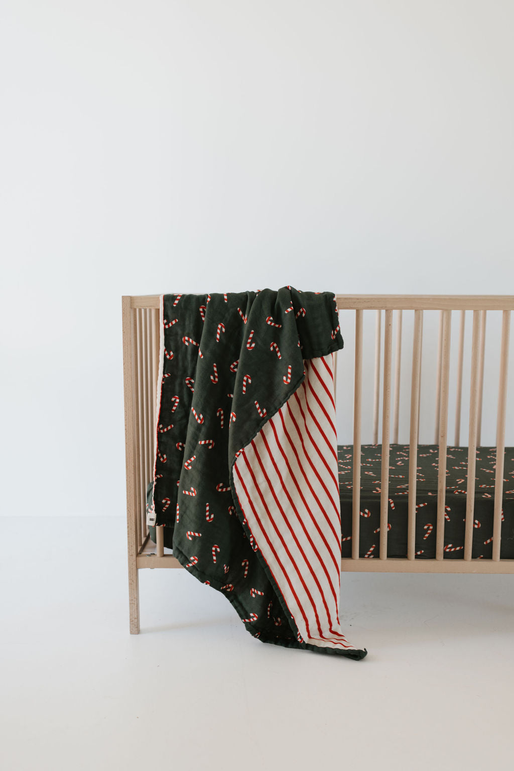 A wooden crib, draped with a festive muslin cotton blanket decorated with candy canes on a dark background, features the Muslin Crib Sheet in the Candy Cane Lane pattern by forever french baby. The blanket's underside is designed with red and white stripes, all set against a minimalistic, white-walled room.