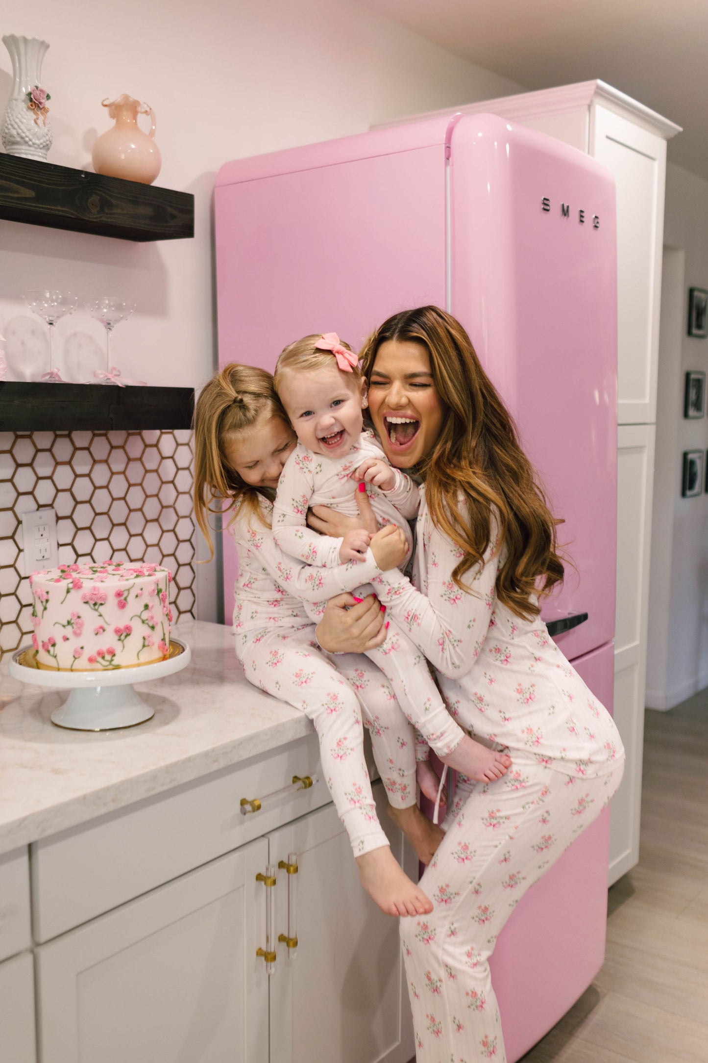 A woman and two young girls in matching Women's Bamboo Pajamas from the Kendy 2 Kendy Floral collection smile joyfully in a kitchen. The woman holds one girl, while another stands on the counter beside a floral cake. Behind them is a pastel pink refrigerator.