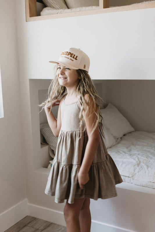 A young girl with long hair stands indoors, smiling while wearing a Vintage Washed Espresso dress by Forever French Baby and a white baseball cap. She is in front of a built-in bed with white beddings in a cozy, well-lit room. Her outfit is part of a matching family set made from organic cotton, adding to the charm of the scene.