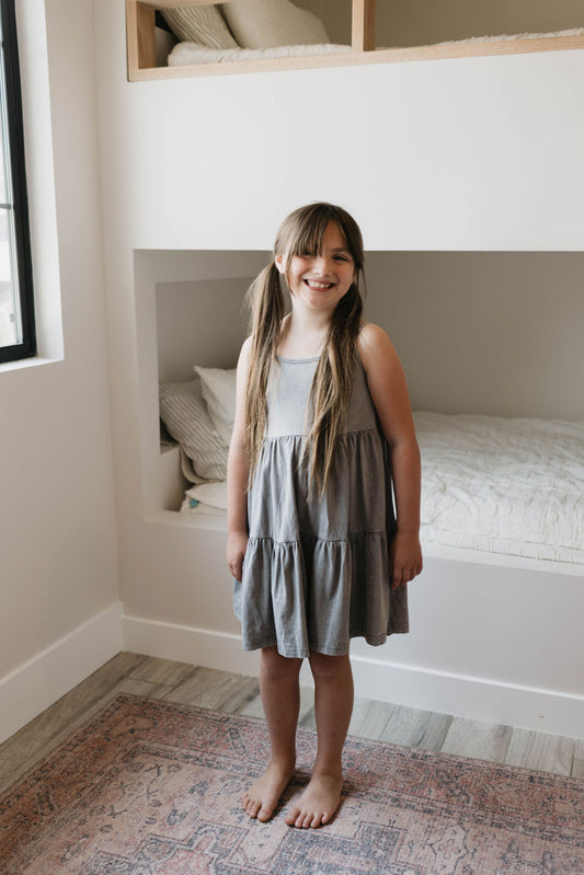 A young girl with long brown hair stands smiling in a cozy bedroom, wearing a sleeveless Children's Dress in Vintage Washed Steel by forever french baby. She is barefoot on a rug, standing in front of a built-in bed adorned with white bedding and shelves above. Natural light streams in from a window on the left.