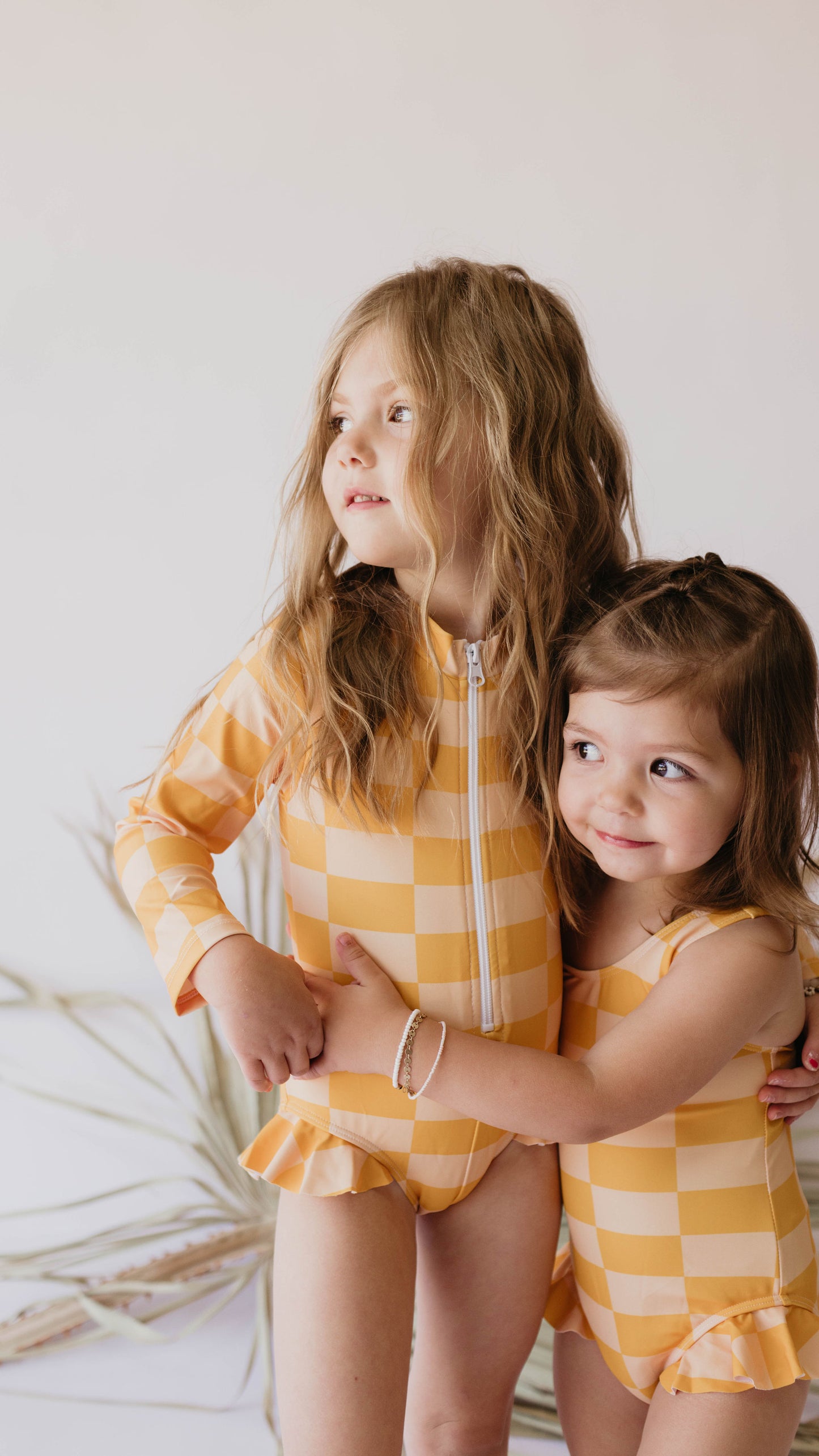 Two young children wearing matching Girl Sleeveless Swimsuits in Sunny Checker print by forever french baby stand together. The older child, with long, wavy hair, looks off to the side, while the younger one, with short hair, smiles and hugs the older sibling. The UPF 50+ fabric ensures they're protected as they play.