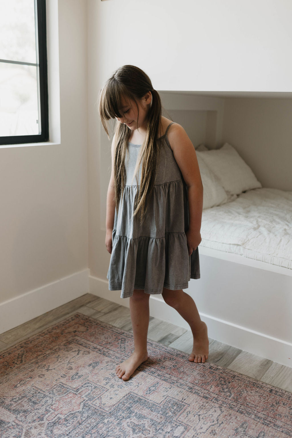 A young girl with long hair stands barefoot on a patterned rug in a minimalistic room. She is wearing the "Children's Dress | Vintage Washed Steel" by forever french baby and looking down with a slight smile. A built-in bed with white bedding is visible in the background.