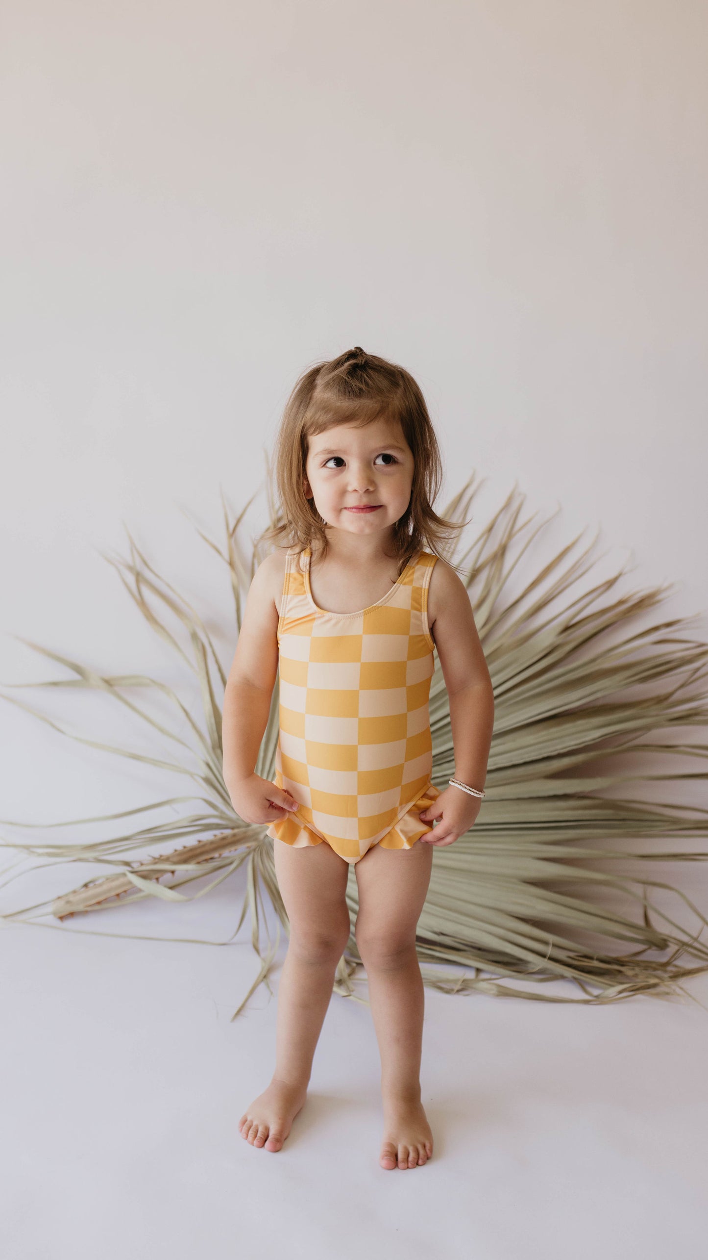 A young child with light brown hair stands barefoot on a white background, wearing the Girl Sleeveless Swimsuit | Sunny Checker from forever french baby. The swimsuit features a cheerful yellow and white checkerboard print. The child smiles slightly and stands in front of a decorative arrangement of dried palm leaves, enjoying the UPF 50+ protection offered by her outfit.