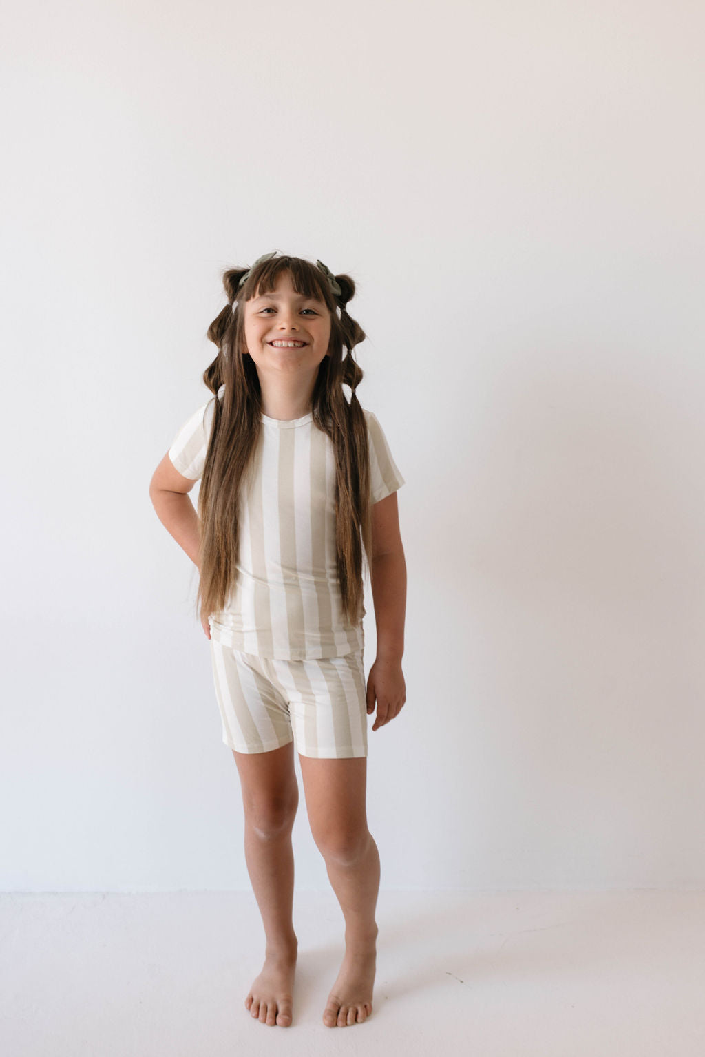 Against a plain white background, a young girl with long brown hair styled in braids and buns stands smiling. She is barefoot and wearing the Bamboo Short Set in Sand Stripe from forever french baby, made from hypoallergenic materials. One hand rests on her hip.