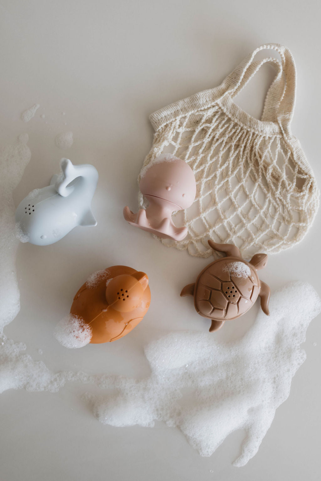 A beige netted bag lies flat on a surface with soap suds scattered around it. Nearby, three mold-resistant bath toys from the "Bath Toys | Ocean Friends Four Pack" by Forever French Baby are arranged: a light blue whale, a pinkish-brown octopus, and a light brown turtle—each adorned with small patches of suds. These charmingly designed toys inspire imaginative play.