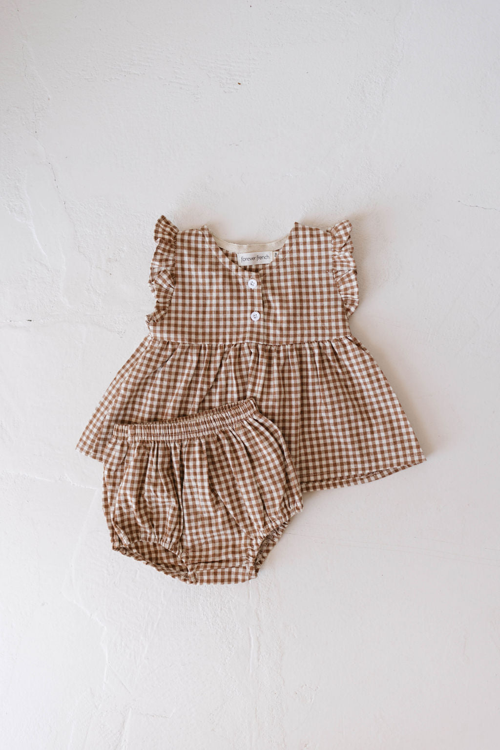 The Gingham Bloomer Set | Honey by Forever French Baby features an infant dress with a delightful gingham print, ruffled sleeves, and matching bloomers, all beautifully displayed on a white background. The dress is designed with front buttons and a gathered waistline, making it perfect for your little one's charming look.