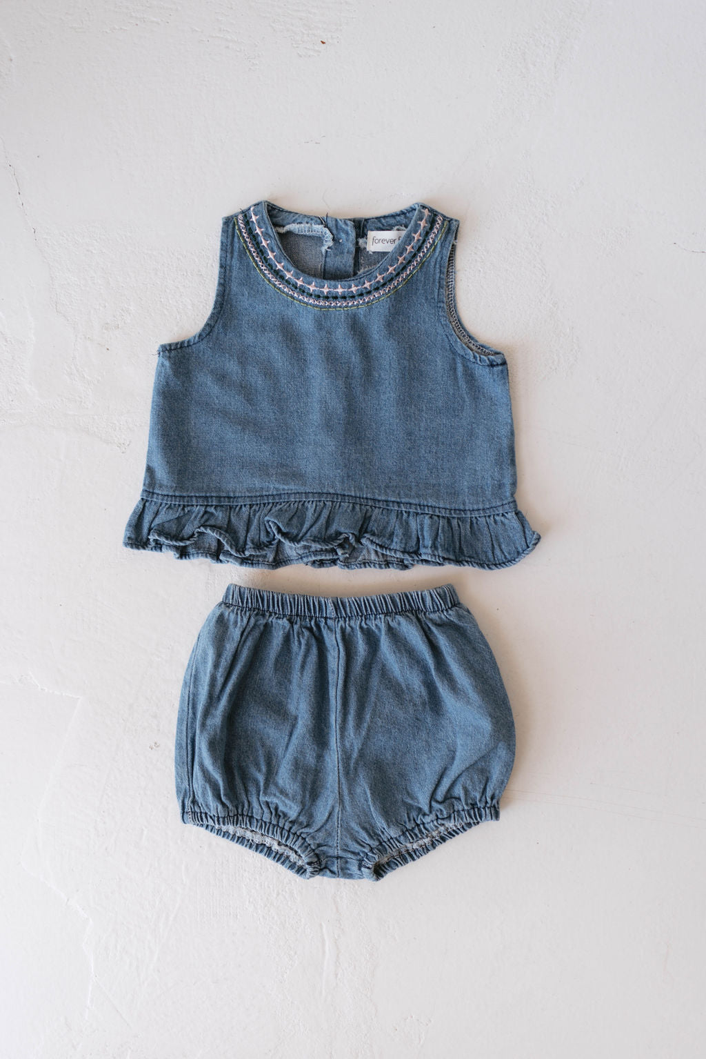 The Denim Bloomer Set | Billie by forever french baby is displayed flat on a white, textured background. This 100% cotton children's outfit features a sleeveless top adorned with embroidered details along the ruffled bottom edge, accompanied by matching bloomers with an elastic waistband and leg openings.