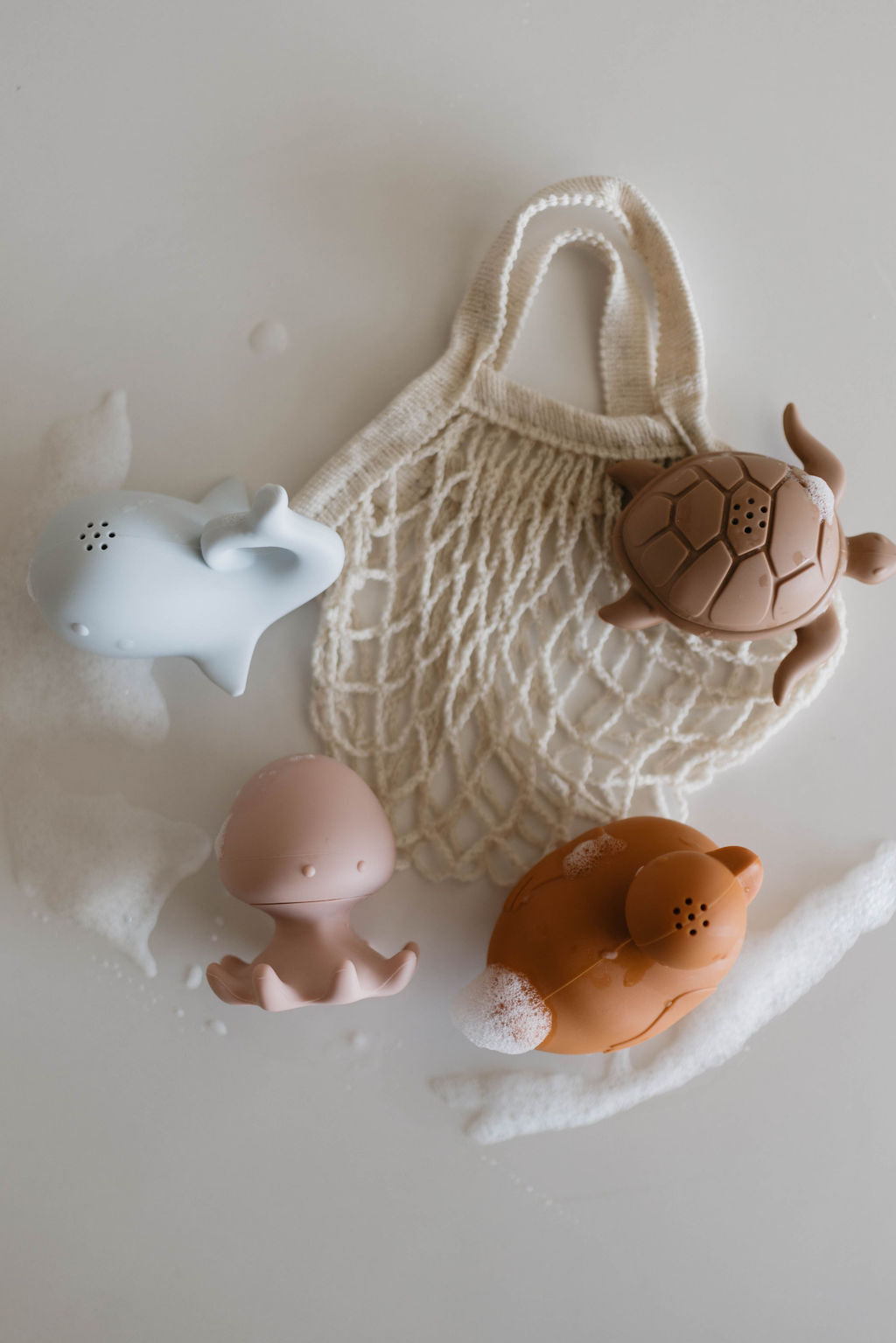 A white net bag on a light surface surrounded by soap suds holds the "Bath Toys | Ocean Friends Four Pack" by forever french baby, featuring four mold-resistant bath toys shaped like sea animals: a gray whale, a brown turtle, a pink squid, and a reddish-orange fish. Perfect for imaginative play with their cute designs.