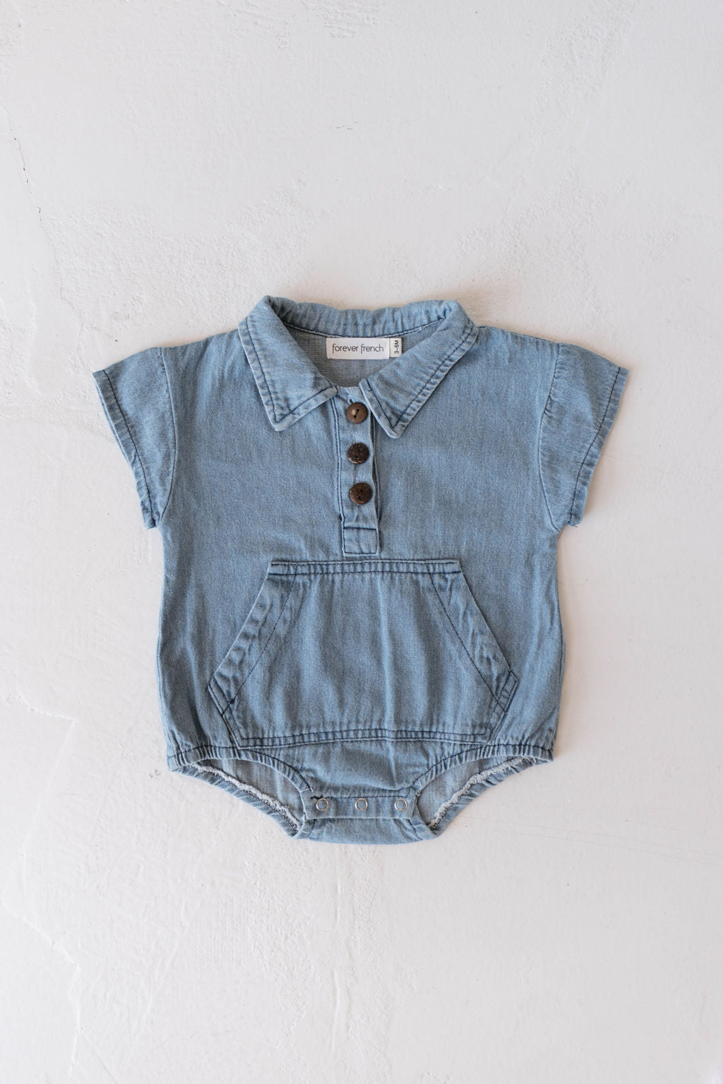 A Denim Romper | Jean from forever french baby is laid flat on a white, textured background. This light blue, short-sleeve baby fashion piece features a collared neck with three brown buttons, a large front pocket, and snap buttons at the bottom for easy diaper changes.