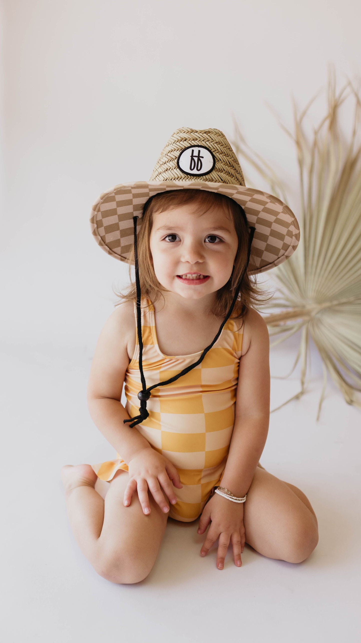 A young child sits on a white surface, smiling at the camera while wearing the Girl Sleeveless Swimsuit in a sunny checkerboard print from forever french baby and a wide-brimmed straw hat with a "bb" logo. A dried palm leaf in the background adds to the radiating summer joy.