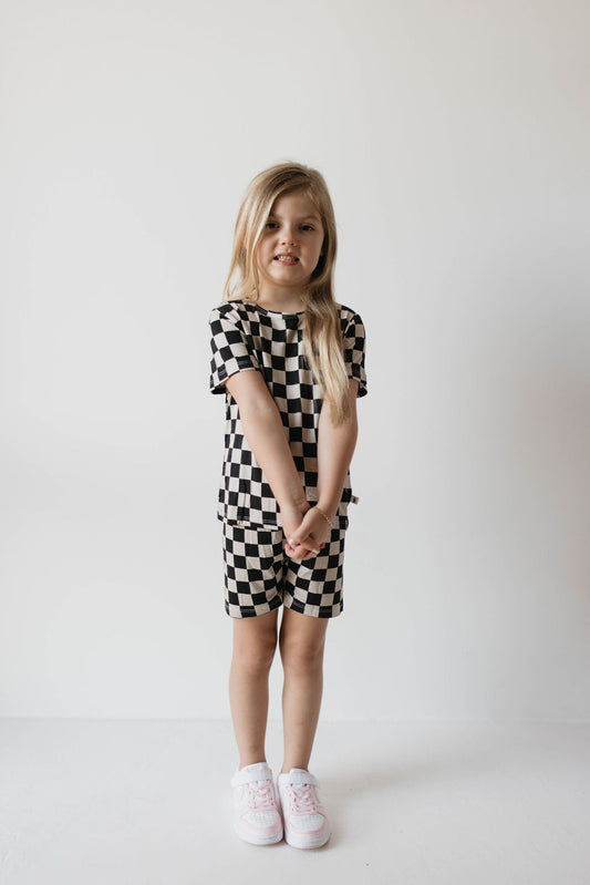 A young girl with long blonde hair stands against a plain white background. She’s dressed in the forever french baby Bamboo Short Set in Black Checkerboard, a breathable outfit featuring a short-sleeved top and matching shorts. White sneakers complete her look as she clasps her hands in front of her.