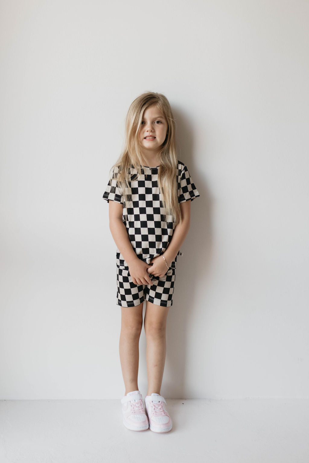 A young girl with long blonde hair stands against a plain white wall. She is wearing the Bamboo Short Set | Black Checkerboard from forever french baby, which includes a short-sleeved top and matching shorts made from breathable fabric. She has on white sneakers and her hands are clasped in front of her.