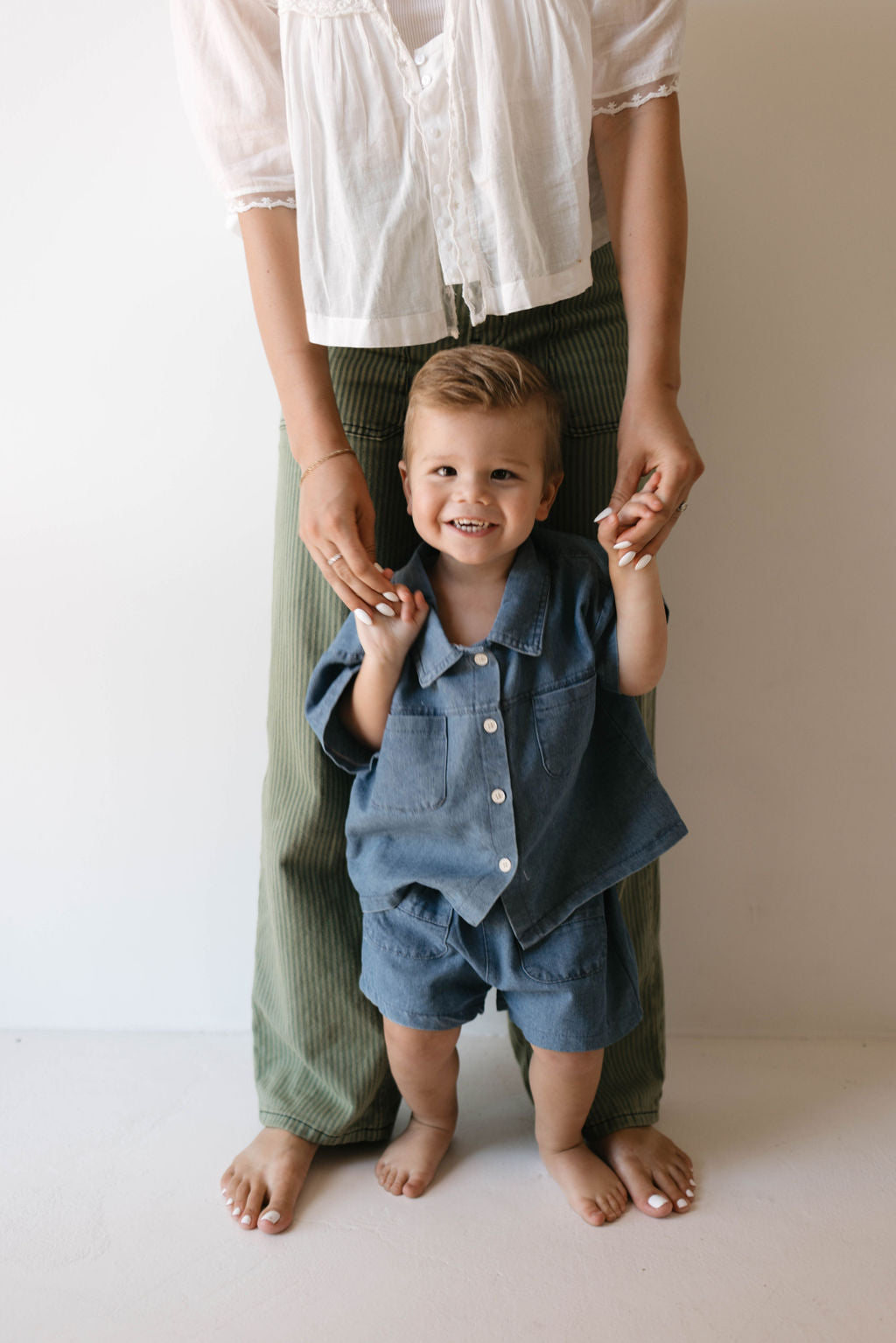 A smiling toddler, dressed in a stylish Denim Short Set | Cash from forever french baby, holds the hands of an adult standing behind him, whose face is out of the frame. The adult is dressed in a white top and green pants. They are standing barefoot on a light floor with a neutral background.