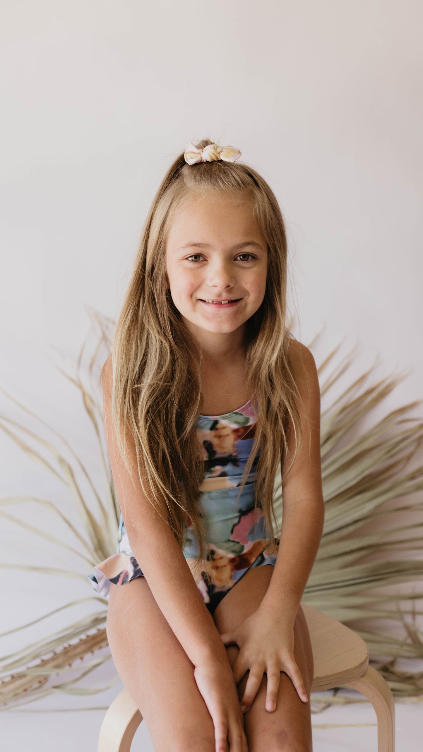 A young girl with long blonde hair, wearing a vibrant Girl One Piece Swimsuit | Charli from forever french baby and a bow in her hair, sits on a wooden stool. She smiles directly at the camera with a neutral background featuring light-toned dried leaves.