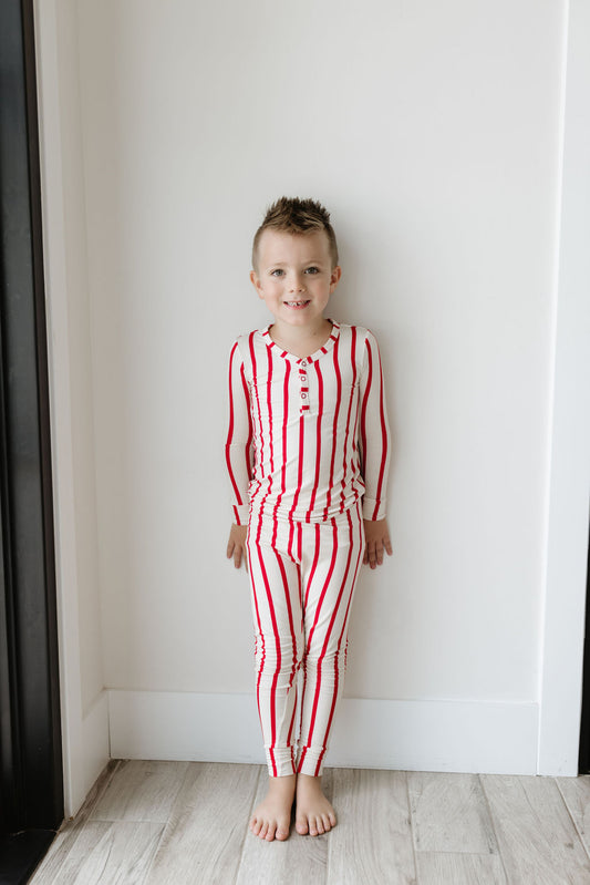 A young child is standing indoors against a white wall, dressed in The Claus Bamboo Two Piece Pajamas from forever french baby. They are barefoot, sporting a joyful smile and short hair. The hypo-allergenic pajamas complement the warm wooden floor as light shines from the left side.