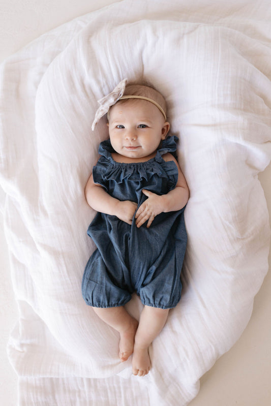 A baby wearing the Forever French Baby 'Betty' denim romper with ruffle tank sleeves and a bow headband lies on a soft white blanket, gently looking up.