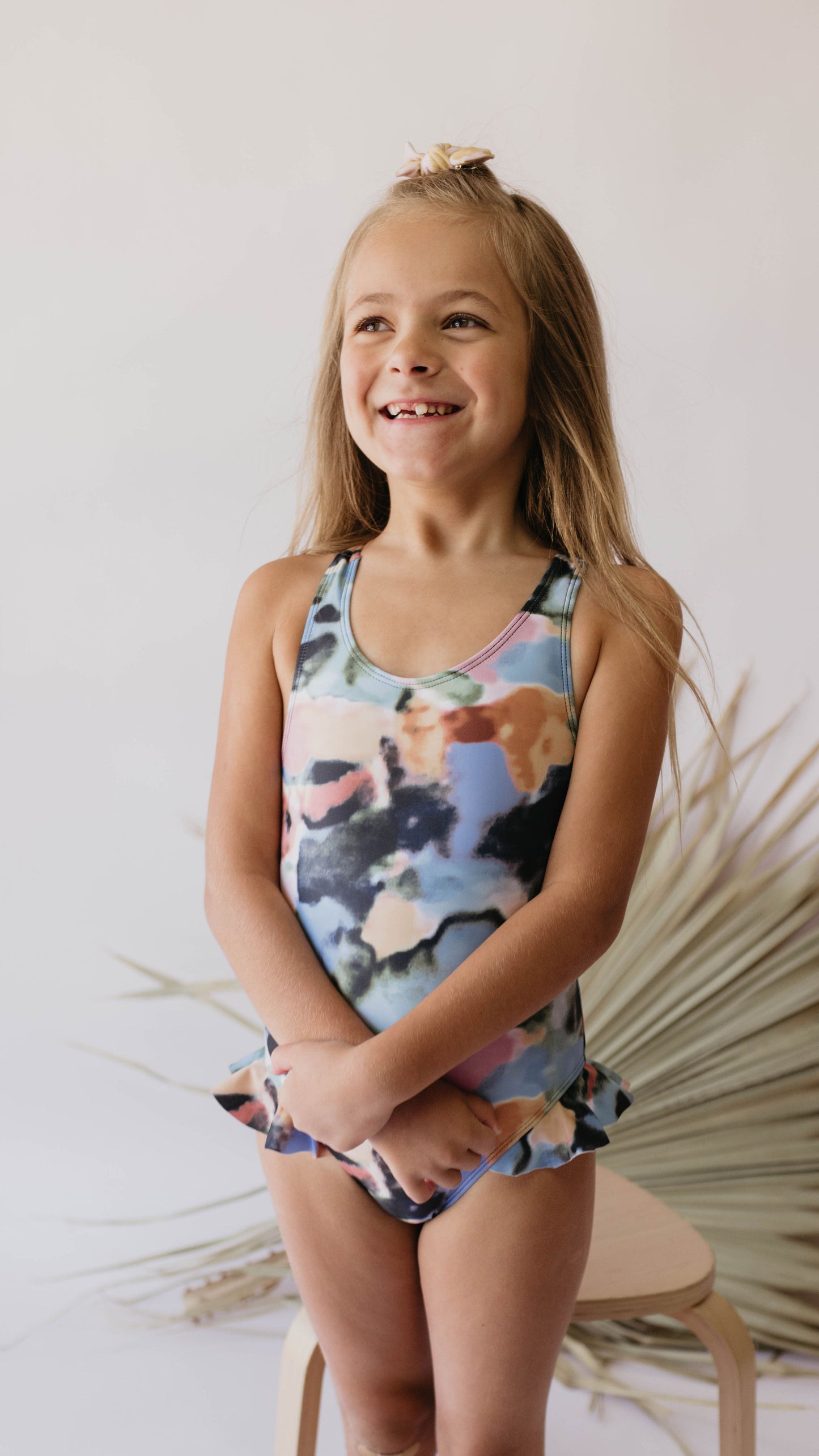 A young girl with long hair stands smiling, hands clasped in front of her. She's wearing the colorful "Girl One Piece Swimsuit | Charli" by forever french baby and standing against a neutral background with decorative dried palm fronds partially visible behind her.