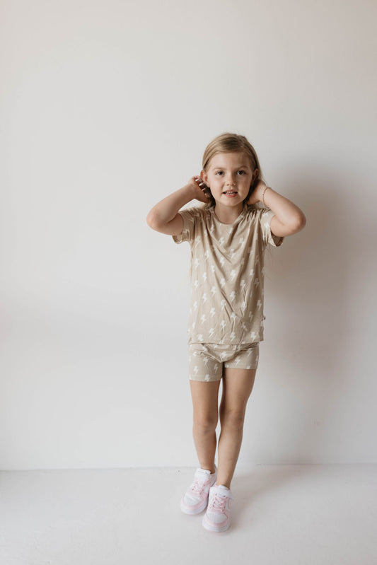 A young girl stands against a plain white background, wearing the Bamboo Short Set | Tan Bolt from forever french baby, featuring a hypo-allergenic beige and white outfit with a nature-inspired pattern. She holds her hands near her ears, with a relaxed and confident expression. She is also wearing pink and white sneakers.