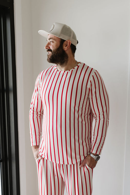 A man stands indoors near a window, wearing the Claus Men's Bamboo Pajamas from forever french baby. The breathable and hypo-allergenic pajamas feature a white and red striped design with matching pants, topped off with a white cap. He has a beard and is smiling while looking to the side.