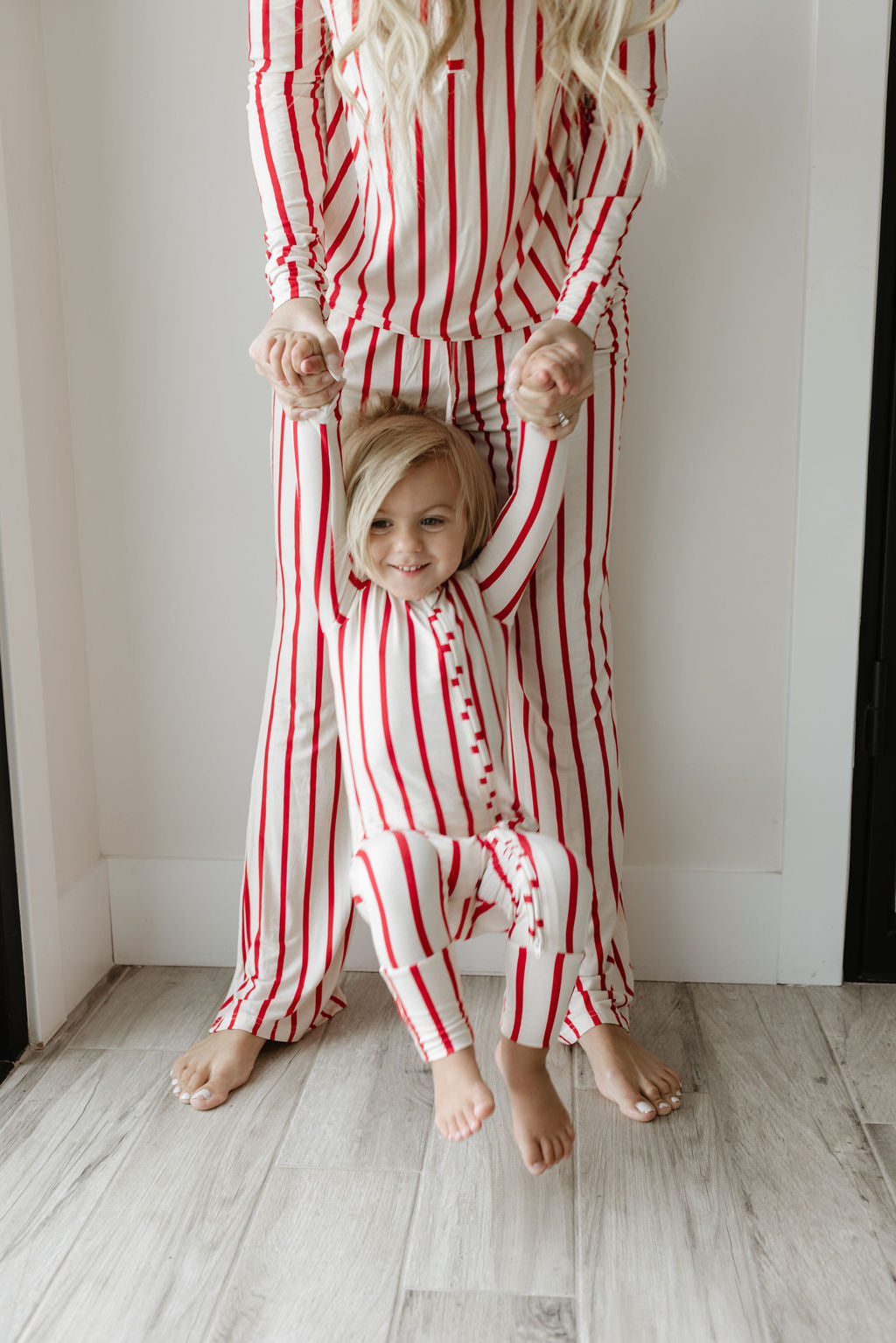 A child wearing the Claus Bamboo Zip Pajamas by forever french baby is playfully lifted by an adult in matching pajamas. The scene unfolds in a bright room with light wooden floors, and the child's joyful expression reflects the comfort of breathable baby clothing.