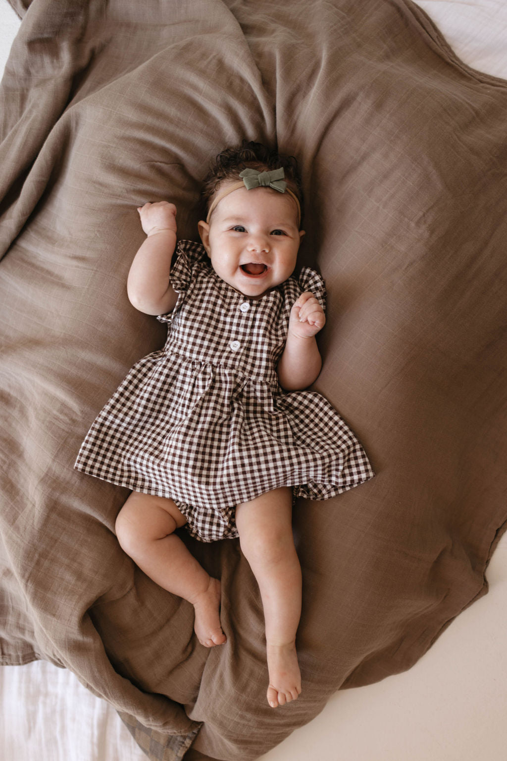 A baby lies on a brown blanket, wearing a Gingham Bloomer Set in Chocolate Chai from forever french baby. The outfit features a charming brown and white gingham print with short sleeves and is made of 100% cotton. A green bow headband adorns the baby's hair as they smile and raise their arms. In the background, a white mattress is partially visible under the blanket.