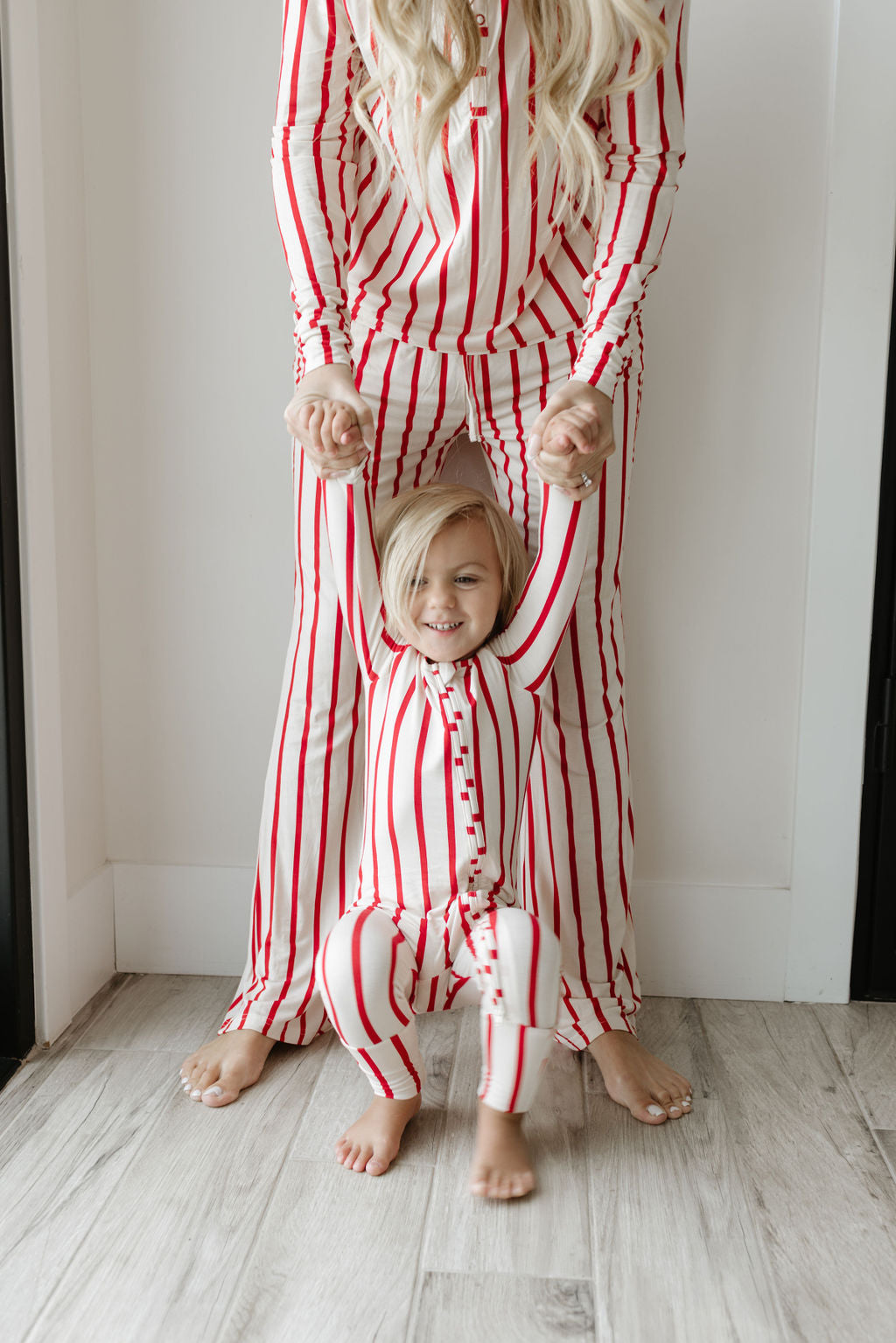 A child wearing forever french baby's Bamboo Zip Pajamas | the Claus, characterized by red and white stripes and hypoallergenic material, is smiling while being supported by their hands in front of an adult in matching sleepwear. They are standing on a wooden floor.