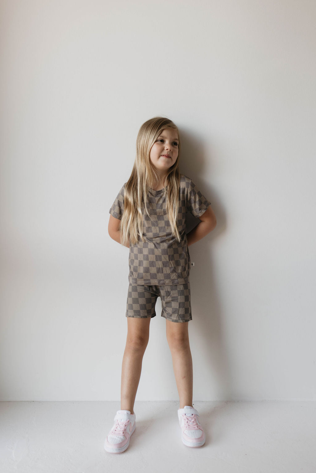 A young girl with long blonde hair stands against a white wall. She is dressed in the Bamboo Short Set | Faded Brown Checker from forever french baby, featuring a breathable T-shirt and shorts. She wears white sneakers with pink accents and has her hands relaxed at her sides, gazing slightly to her left.