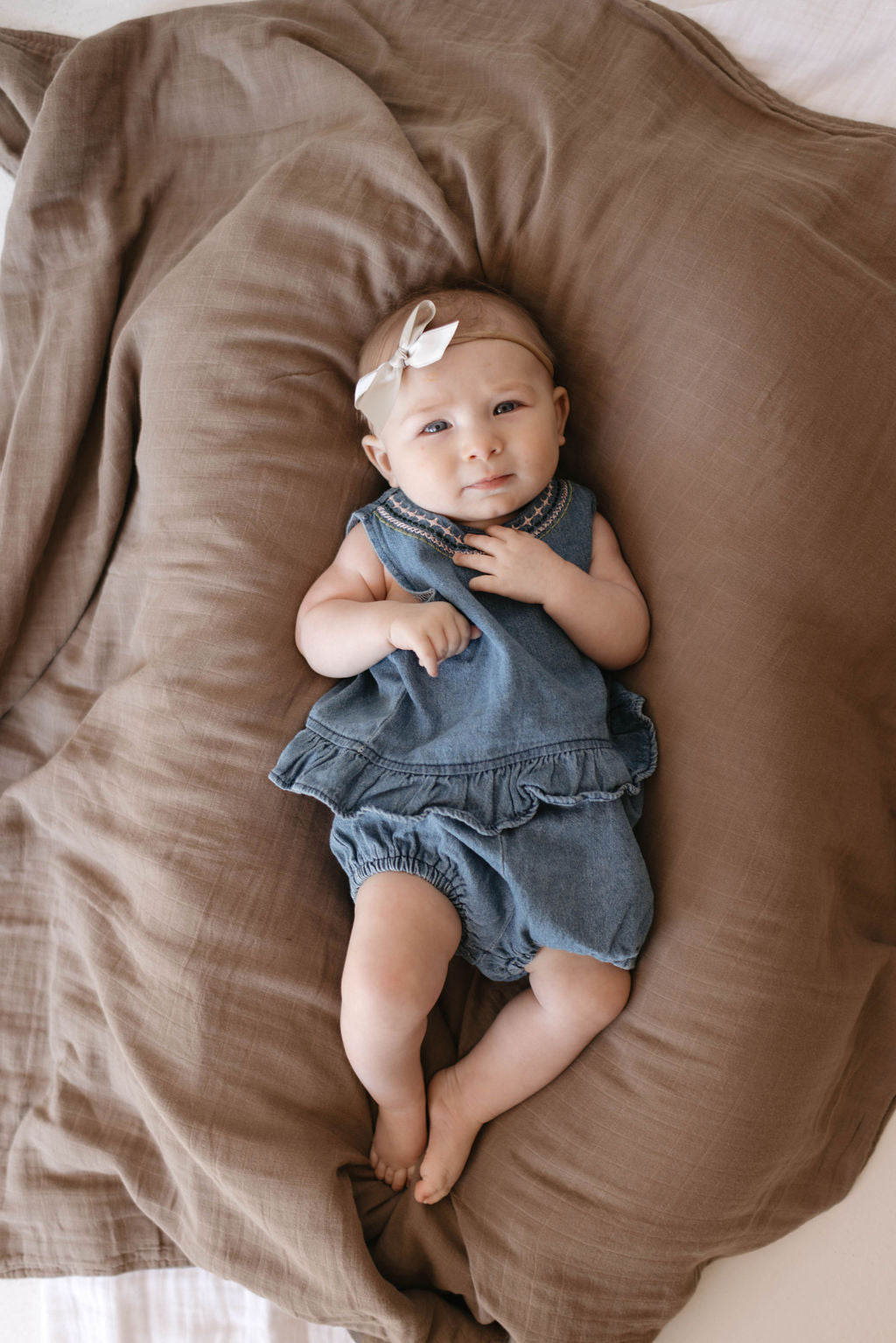 A baby dressed in the Denim Bloomer Set from forever french baby, featuring ruffled edges and embroidered details, lies on a large brown cushion, complemented by a beige headband with a small bow. The baby looks up with a slight smile, surrounded by soft, neutral-toned bedding.