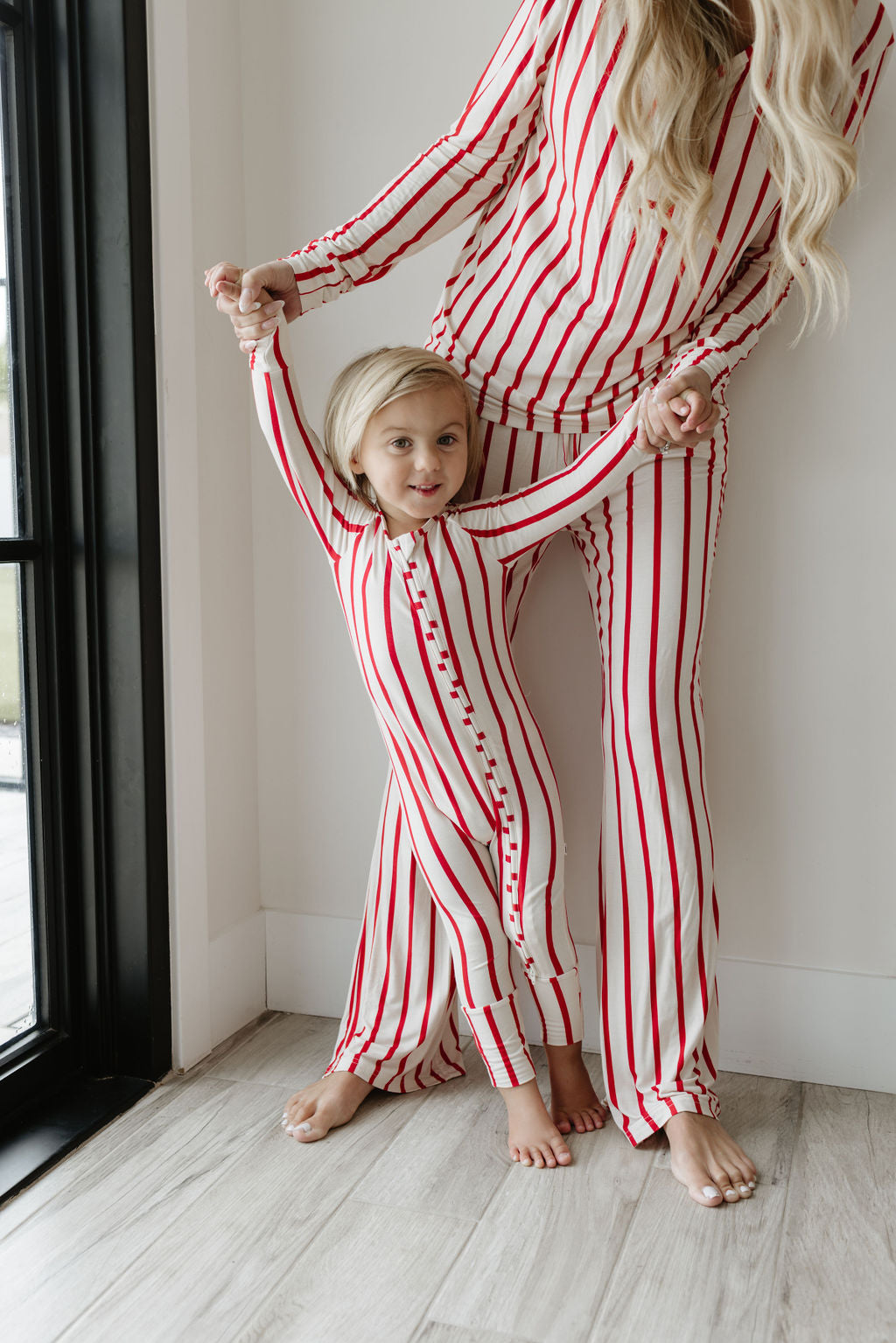 An adult and child, smiling warmly, stand barefoot on a wooden floor near a window, dressed in matching red-striped white Bamboo Zip Pajamas by forever french baby. The adult holds the child’s hands gently.