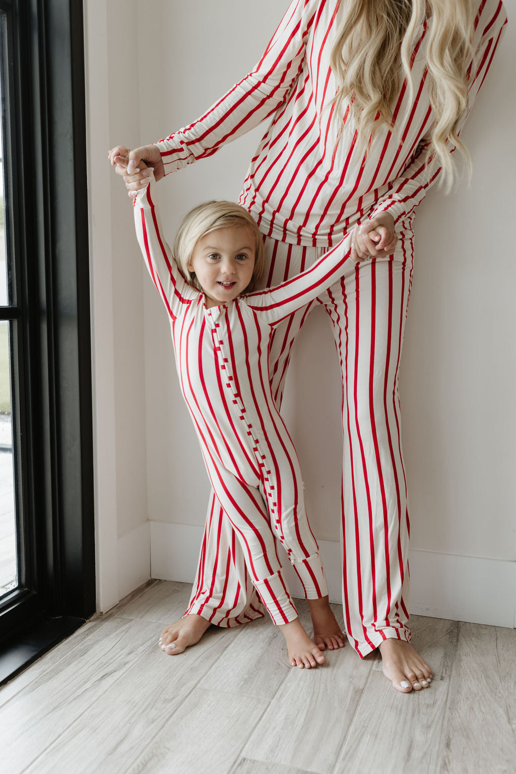 A parent and child, dressed in matching Bamboo Zip Pajamas from the Claus collection by forever french baby, hold hands and smile near a window. The child stands in front looking cheerful in their hypoallergenic sleepwear. Both are barefoot on the wooden floor.