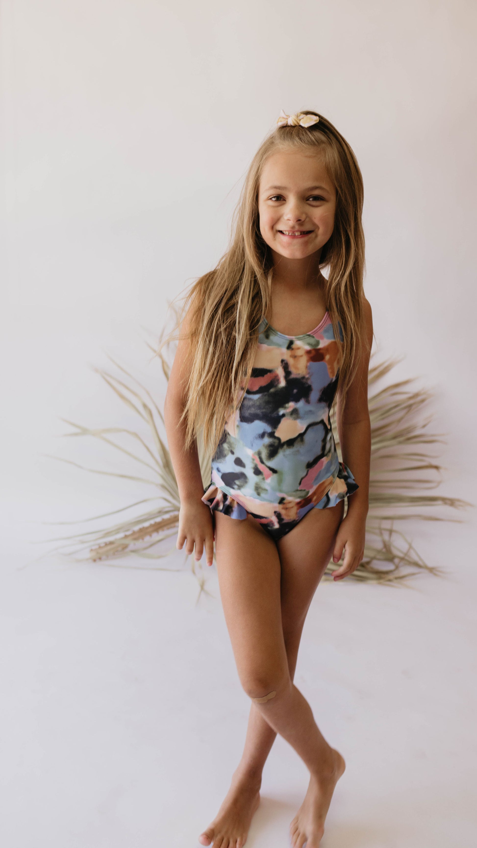 A young girl with long blonde hair stands smiling, wearing a colorful "Girl One Piece Swimsuit | Charli" by forever french baby. She is barefoot and posing in front of a neutral background with dried palm leaves, promoting Autism awareness.