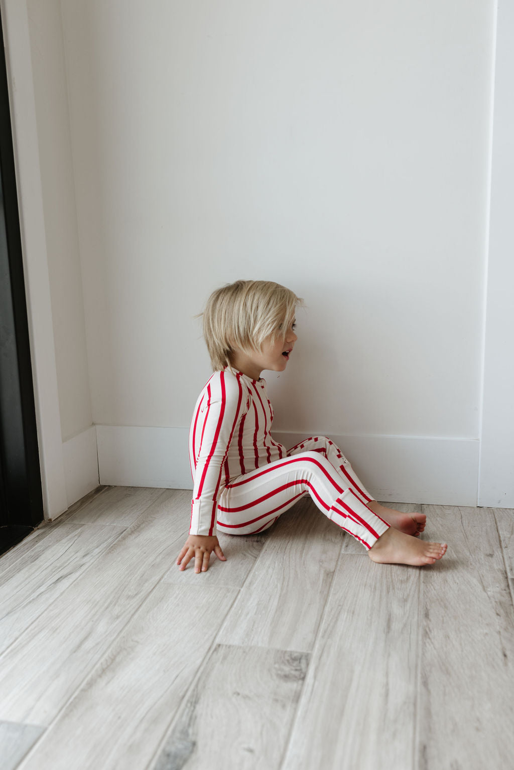 A young child with blond hair sits on the floor against a white wall, wearing the Bamboo Zip Pajamas | the Claus by forever french baby. The hypoallergenic, red and white striped sleepwear ensures comfort as the child, barefoot and carefree, gazes slightly to the side on light wood-patterned flooring.