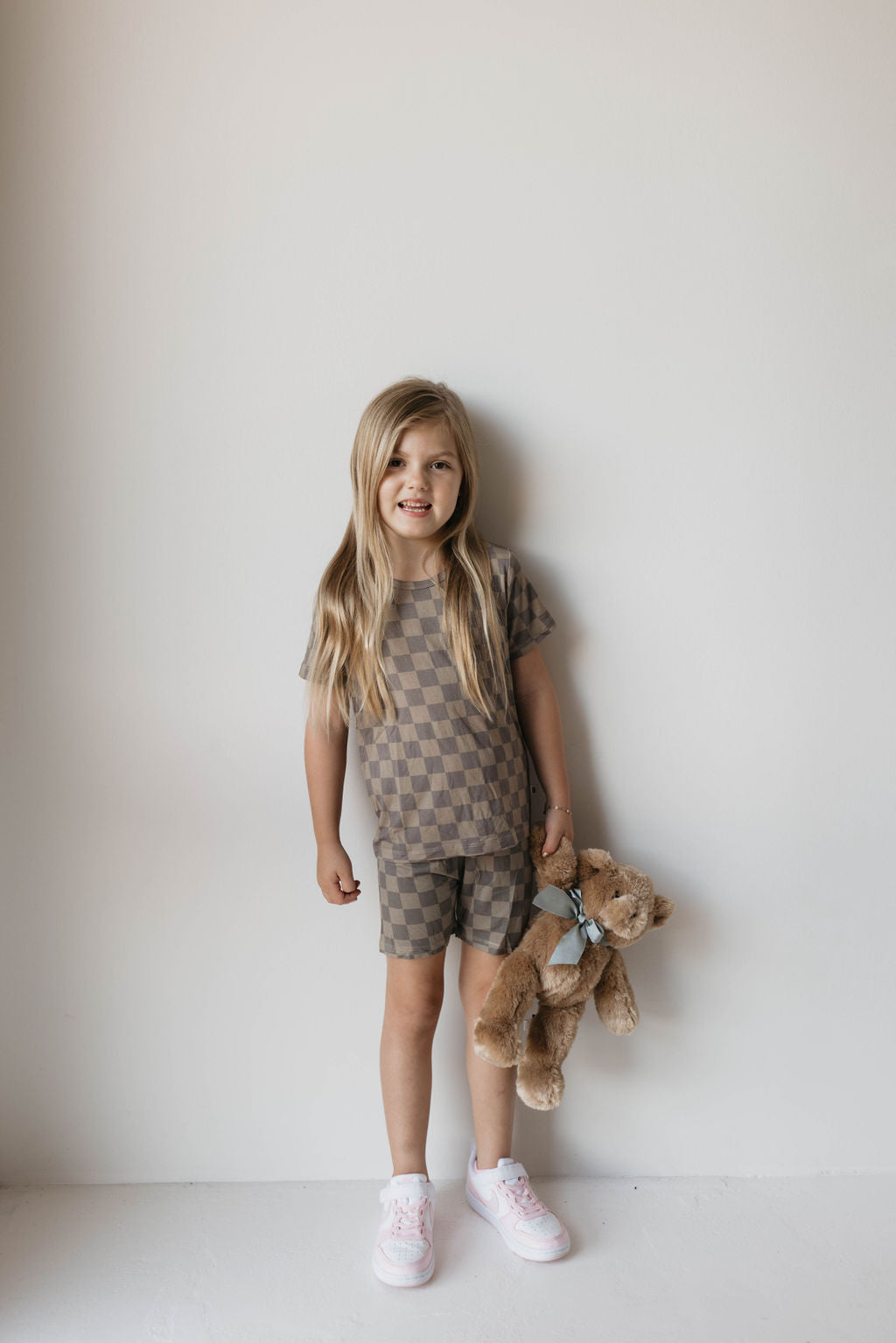 A young girl with long blond hair stands against a plain white wall. She is wearing the Bamboo Short Set in Faded Brown Checker by forever french baby, known for its hypo-allergenic fabric, and light pink sneakers. She holds a brown teddy bear in her right hand and smiles slightly.