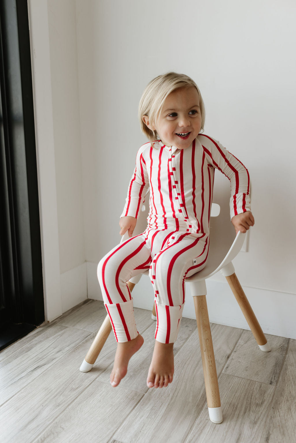 A child with long blond hair is sitting on a small chair indoors, wearing the Bamboo Zip Pajamas | the Claus from forever french baby. The pajamas feature breathable white fabric with red stripes. The child is smiling and looking slightly to the side in a light-colored room with wooden flooring.