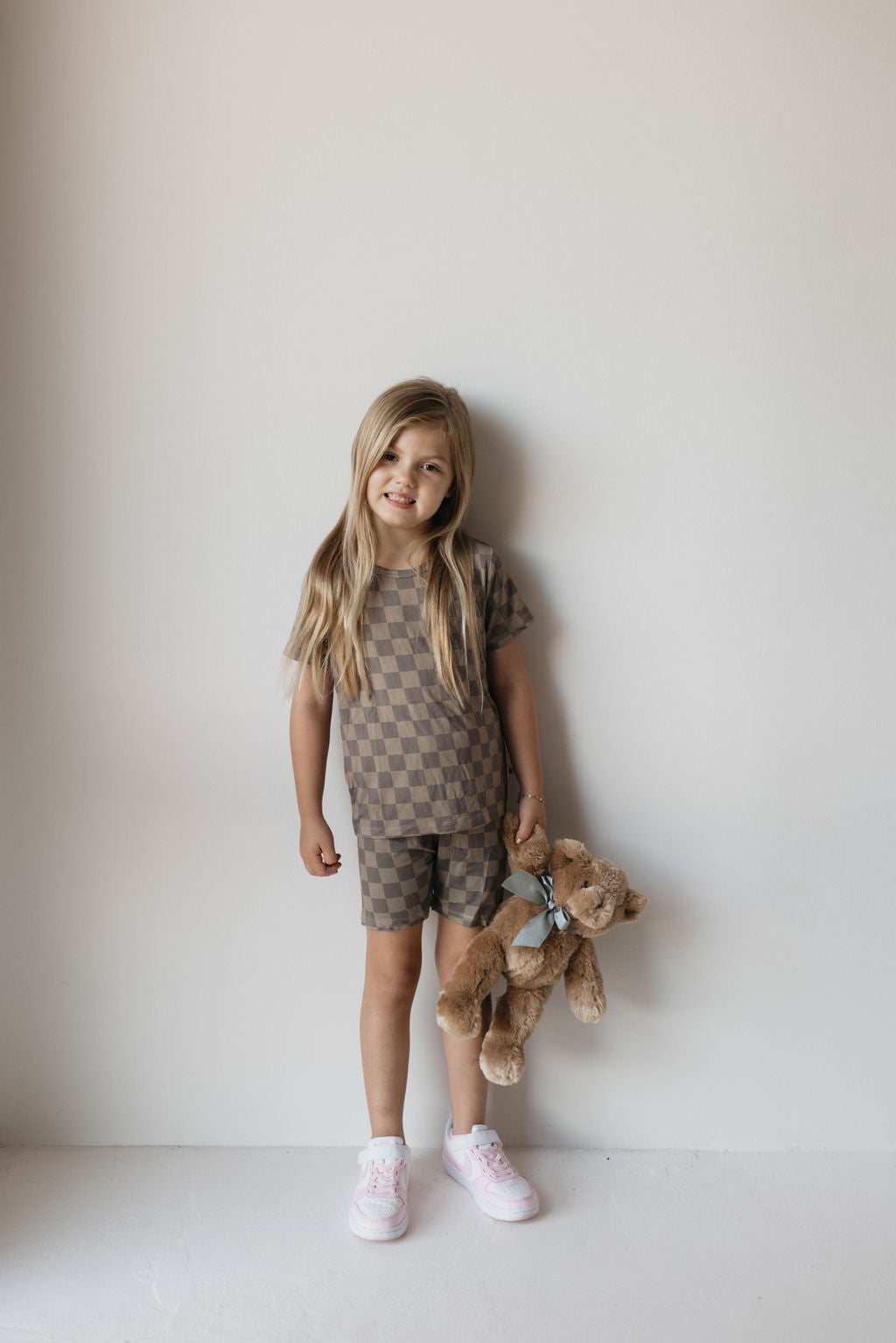 A young child with long hair stands against a plain white wall, smiling and wearing the Bamboo Short Set | Faded Brown Checker from forever french baby. The set includes a checkered, short-sleeved shirt and matching shorts made from breathable material. The child is holding a brown stuffed toy and wearing pink sneakers.